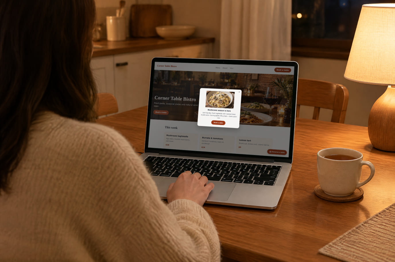 A guest at her kitchen table in the evening opens the bistro website on a laptop, with the promo popup on screen